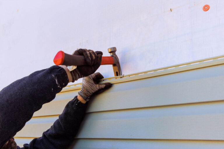 siding repair-close up of a man wearing safety gloves using a hammer to remove old siding materials from a house